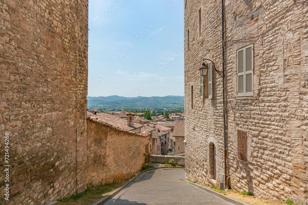 Blue sky in view between two tall stone walls and cobbled street leading downhill Gubbio, medieval town.