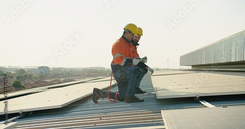 Two technicians in safety harnesses are working on the installation of solar panels on a sunny rooftop against a clear sky background