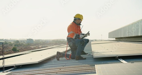 Two technicians in safety harnesses are working on the installation of solar panels on a sunny rooftop against a clear sky background
