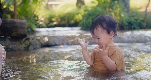 A curious toddler in a brown shirt joyfully explores the sensations of splashing water experiences the joy of water play in a shallow stream, surrounded by lush vegetation