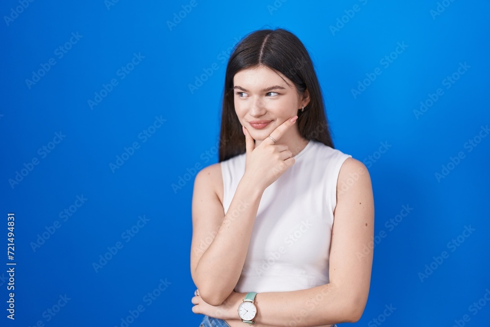 Young caucasian woman standing over blue background looking confident at the camera smiling with crossed arms and hand raised on chin. thinking positive. Young caucasian woman standing over blue background looking confident at the camera smiling with crossed arms and hand raised on chin. thinking positive.