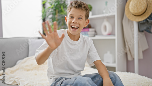 Adorable blond boy full of joy and confidence, relaxingly sitting on the living room sofa at home but cheerfully saying hello with his hand, grinning from ear to ear photo