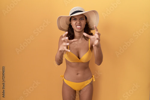 Young hispanic woman wearing bikini and summer hat shouting frustrated with rage  hands trying to strangle  yelling mad