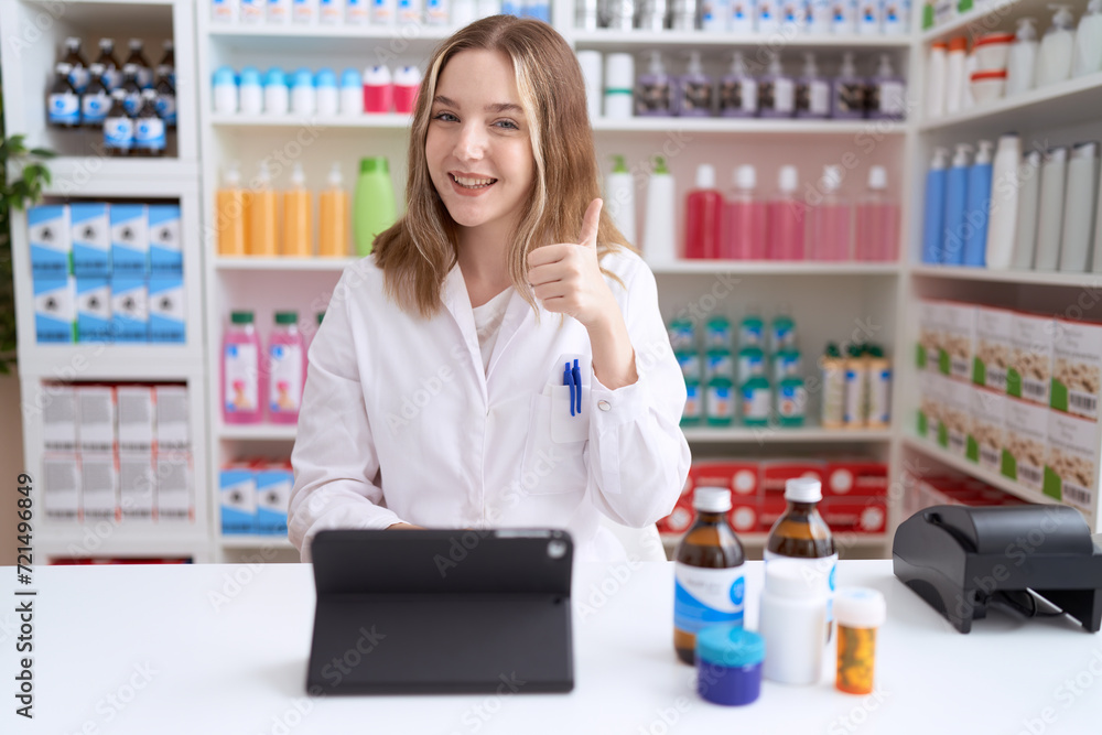 Young caucasian woman working at pharmacy drugstore using tablet smiling happy and positive, thumb up doing excellent and approval sign
