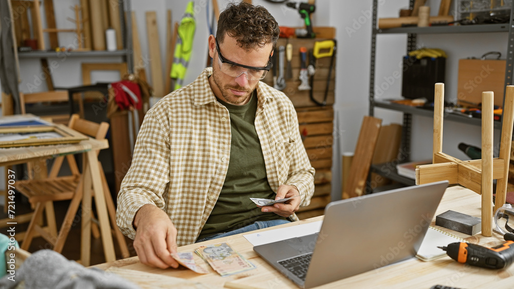 Bearded man examines money in a carpentry workshop with a laptop open nearby. Bearded man examines money in a carpentry workshop with a laptop open nearby.