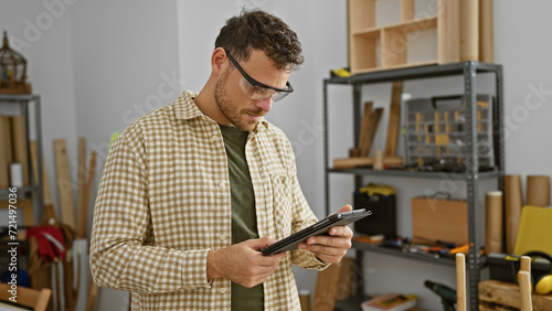 A focused young man with a beard checks a tablet in a well-equipped carpentry workshop.