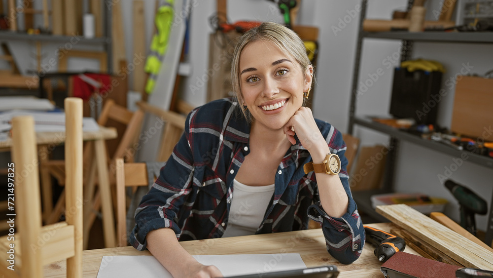 Smiling woman with blonde hair in a plaid shirt posing in a carpentry workshop surrounded by woodworking tools.