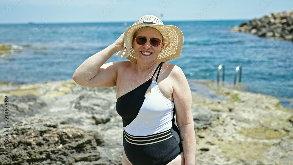 Middle age grey-haired woman tourist wearing swimsuit and summer hat smiling at the beach Middle age grey-haired woman tourist wearing swimsuit and summer hat smiling at the beach