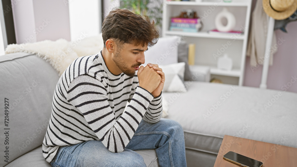 Worried young arab man with a beard, sitting on his living room sofa at home, seriously expressing doubt and sadness as he nervously ponders a problem indoors.