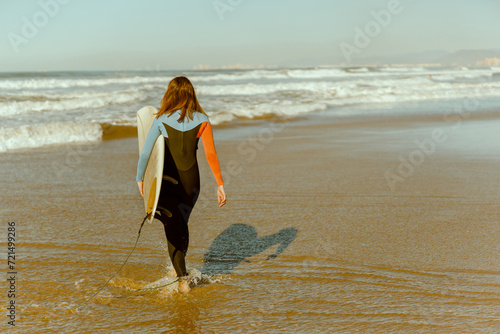Back view of female surfer in wetsuit with his surfboard entering the sea for riding on waves