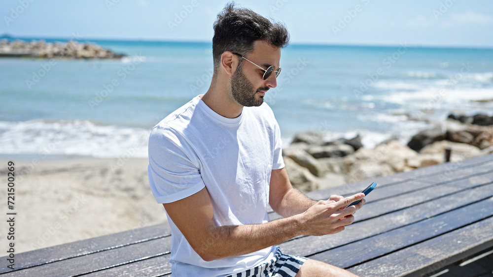 Young hispanic man using smartphone with serious expression at seaside