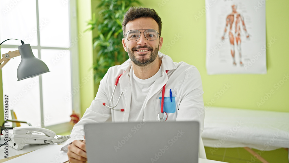 Young hispanic man doctor using laptop wearing glasses smiling at clinic Young hispanic man doctor using laptop wearing glasses smiling at clinic