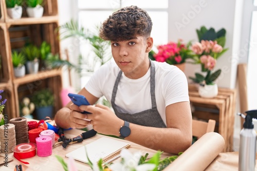 Young hispanic teenager florist using smartphone sitting on table at flower shop