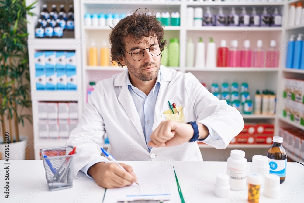 Hispanic young man working at pharmacy drugstore checking the time on wrist watch, relaxed and confident