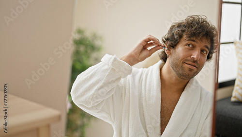 Young hispanic man wearing bathrobe cleaning ear with cotton swab looking on mirror at home