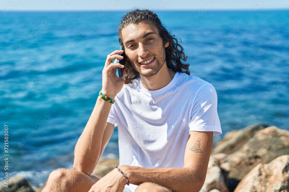 Young hispanic man talking on smartphone sitting on rock at seaside