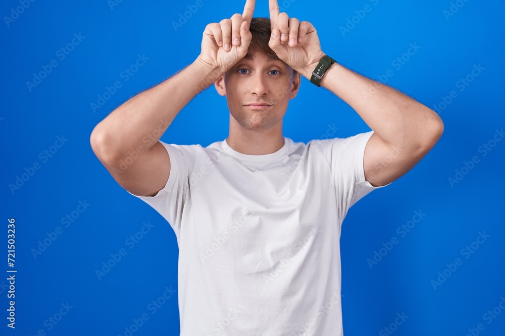 Naklejka premium Caucasian blond man standing over blue background doing funny gesture with finger over head as bull horns
