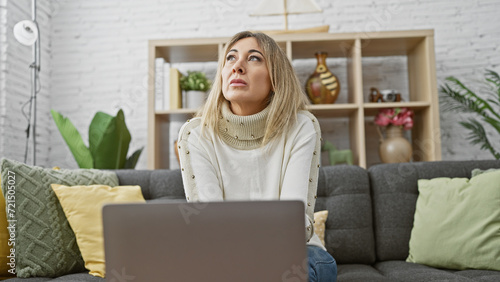 Pensive blonde woman sitting on a sofa at home, looking upwards, in a cozy living room setting.