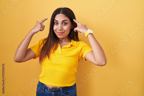 Young arab woman standing over yellow background smiling pointing to head with both hands finger, great idea or thought, good memory