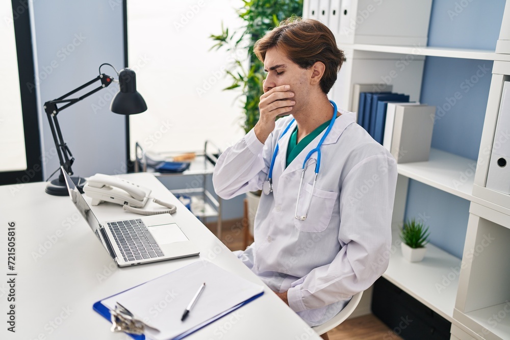 Young caucasian man doctor sitting on table yawning at clinic