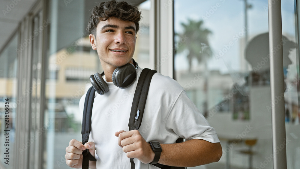 Fototapeta premium Cheerful young hispanic male student standing outdoors enjoying campus life, confidently wearing headphones and backpack, radiating positive energy and joy at the university.
