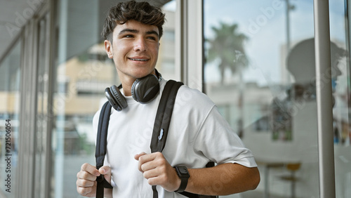 Cheerful young hispanic male student standing outdoors enjoying campus life, confidently wearing headphones and backpack, radiating positive energy and joy at the university. photo