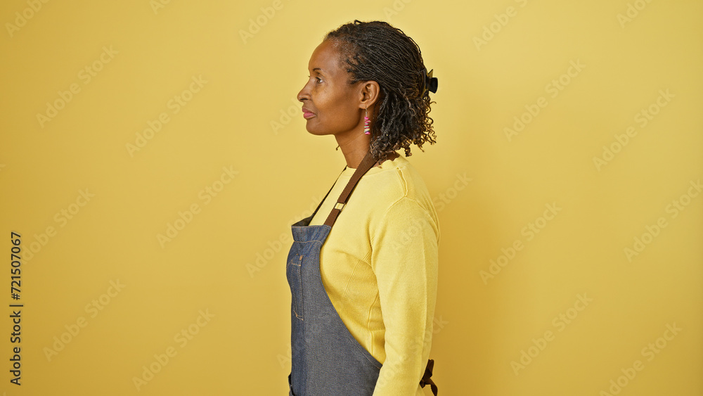 Fototapeta premium Profile of a mature african woman wearing a yellow sweater and denim apron against an isolated yellow background