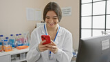 Hispanic woman in laboratory smiles while using smartphone, capturing a moment of modern healthcare professionalism.