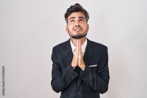 Obraz na płótnie Young hispanic man with tattoos wearing business suit and tie begging and praying with hands together with hope expression on face very emotional and worried