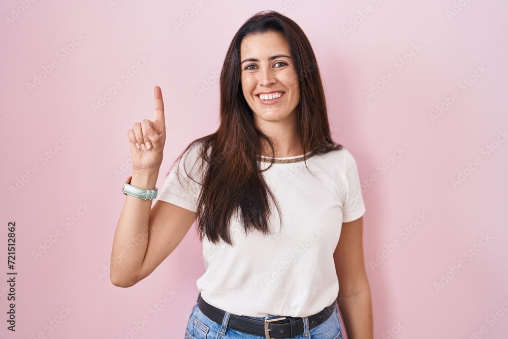 Young brunette woman standing over pink background showing and pointing up with finger number one while smiling confident and happy. Young brunette woman standing over pink background showing and pointing up with finger number one while smiling confident and happy.