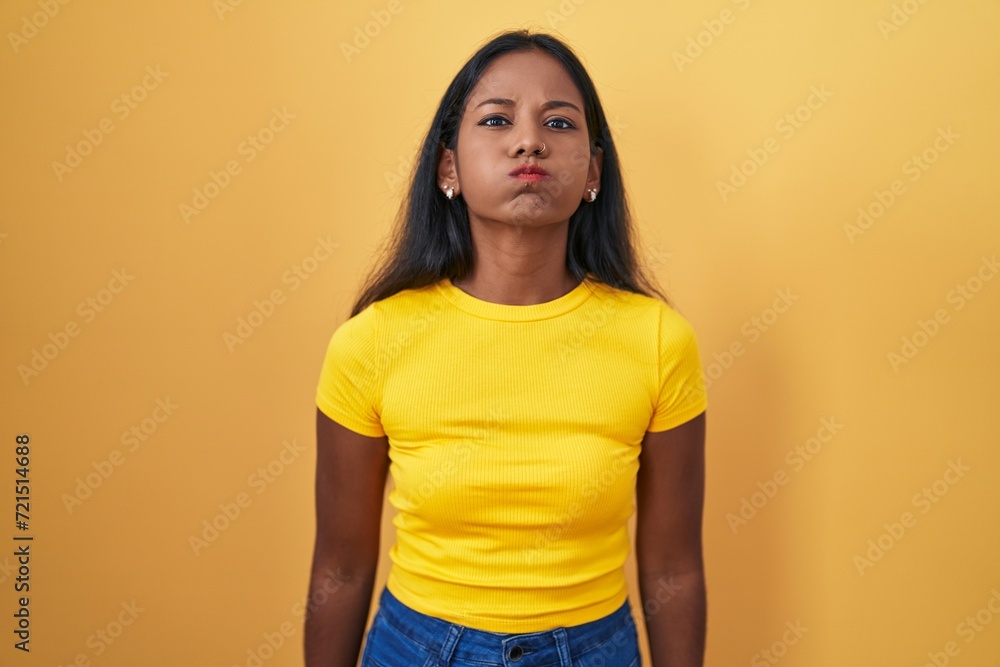 Obraz premium Young indian woman standing over yellow background puffing cheeks with funny face. mouth inflated with air, crazy expression.