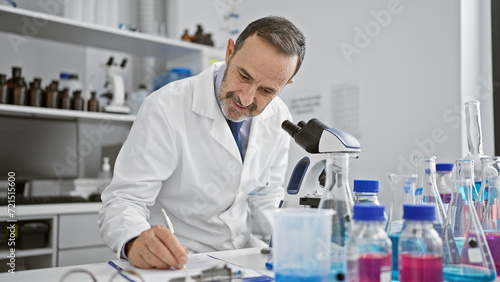 Middle age man with grey hair scientist using microscope writing on clipboard at laboratory