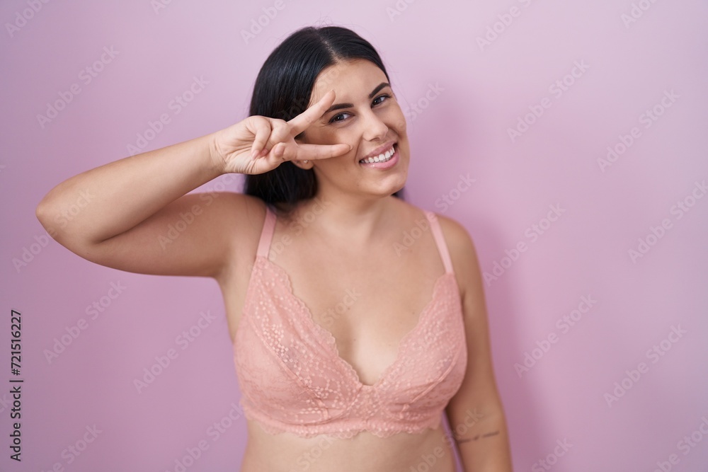Young hispanic woman wearing pink bra doing peace symbol with fingers over face, smiling cheerful showing victory Young hispanic woman wearing pink bra doing peace symbol with fingers over face, smiling cheerful showing victory