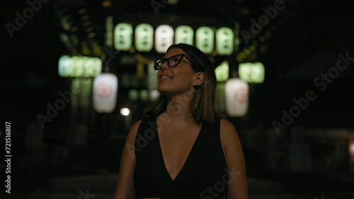 Cheerful beautiful hispanic woman in glasses enjoying kyoto's nightlife, looking around and smiling confidently on a lighted japanese street