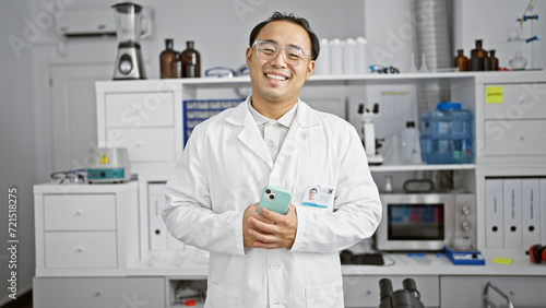 Fototapeta Naklejka Na Ścianę i Meble -  Smiling young chinese scientist confidently navigates his smartphone in bustling lab amidst test tubes and high-tech research