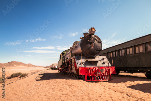 an old black, green and red vintage steam train rests on the tracks in a wild and sunny desert environment, with carriages in the background