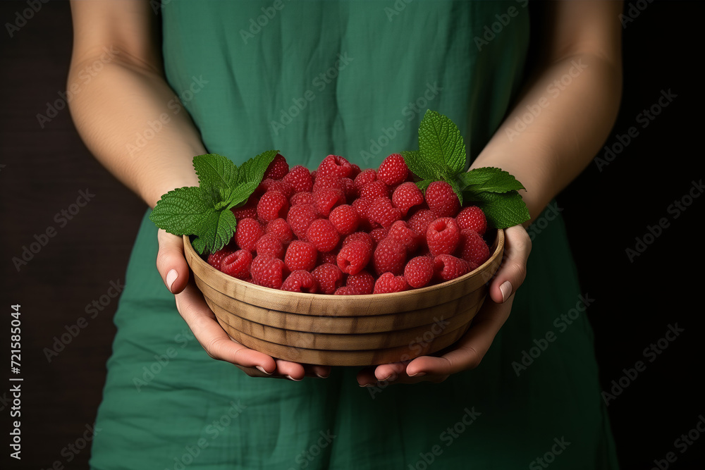 Ripe fresh healthy raspberries with leaves in a wooden bowl, plate in the hands of a girl in a green linen dress on a dark background Ripe fresh healthy raspberries with leaves in a wooden bowl, plate in the hands of a girl in a green linen dress on a dark background