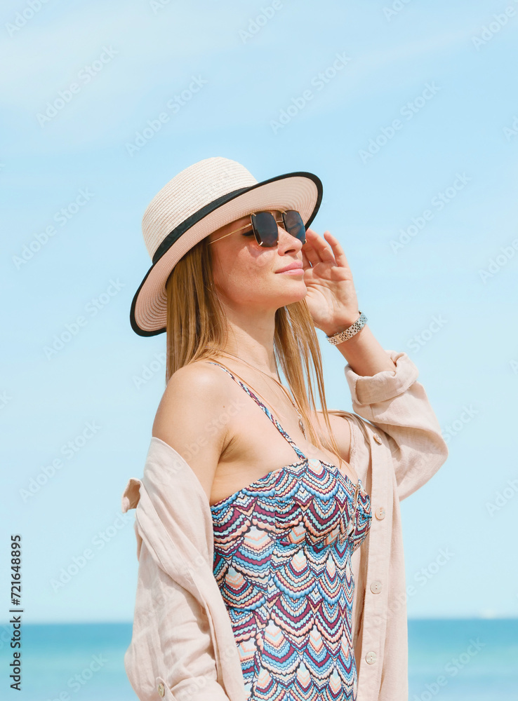 Young woman in a swimsuit, shirt, sunglasses and hat posing gracefully on a sandy beach against a blue sky