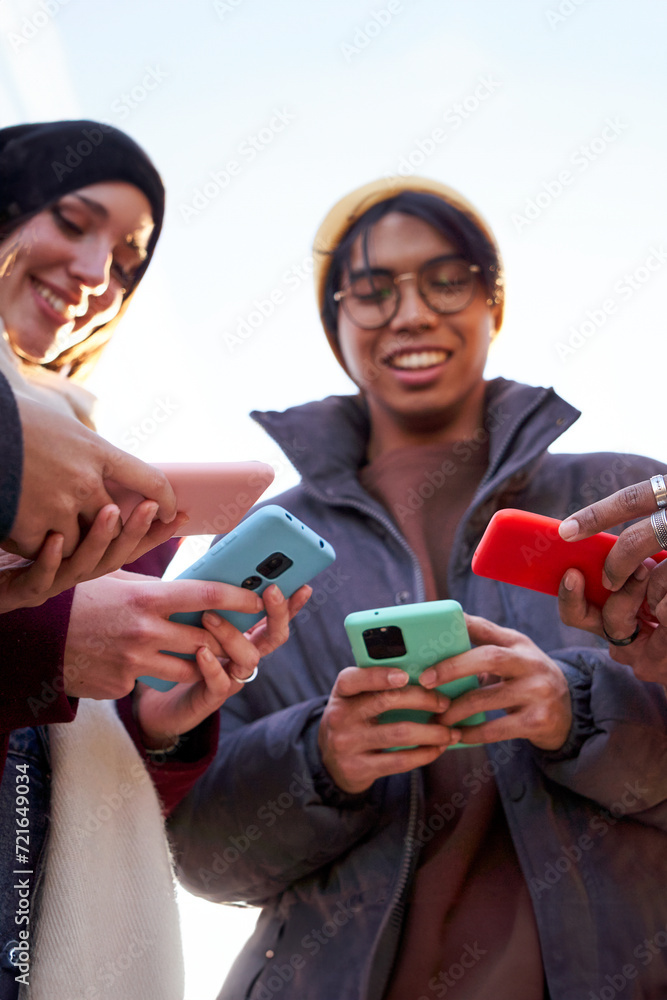 Obraz premium Multiracial friends standing in circle using phone outdoors. Low angle view, vertical image with copy space. Concept of technology addiction.