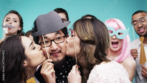 Happy people, party and kiss man on cheek, moustache and event celebration with excited friends. Dancing, holiday and photobooth with men, woman and social gathering on studio background for fun photo
