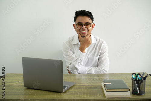 young asian businessman in a workplace smiling with holding chin and looking at camera, wearing white shirt with glasses isolated