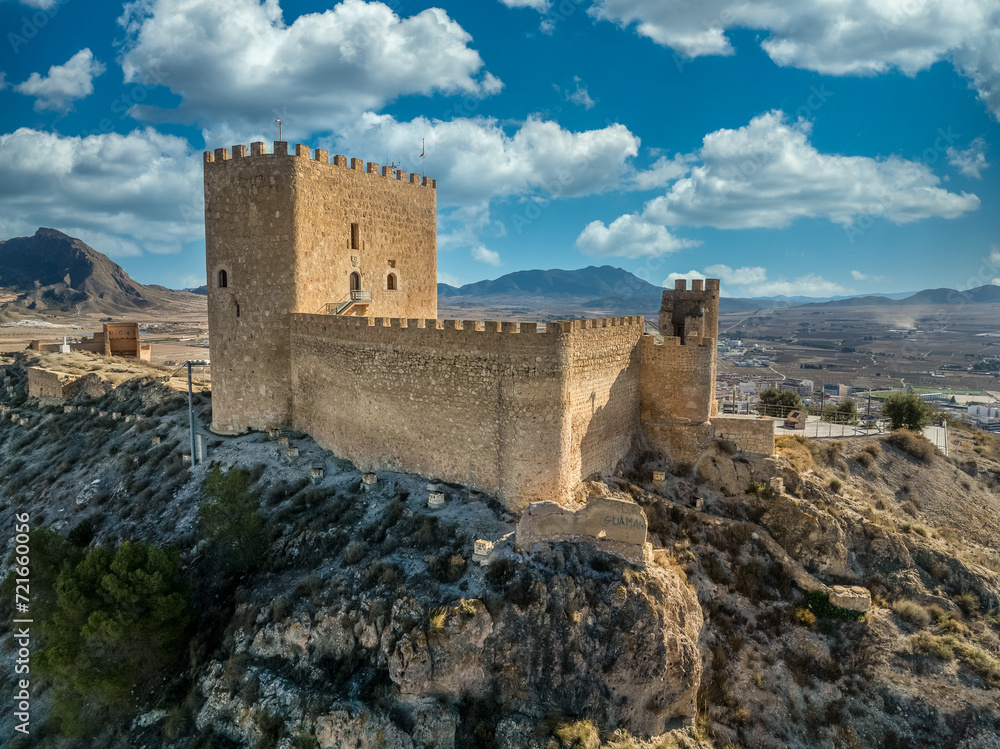 Aerial view of Jumilla medieval castle in Murcia Spain, on a hilltop, imposing irregular shape keep with four floors, crenelated battlements cloudy blue sky background