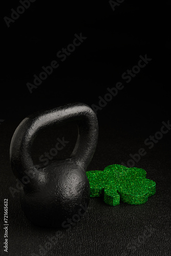 Fototapeta Naklejka Na Ścianę i Meble -  Holiday fitness and Happy St. Patick’s Day, green glitter shamrock shape with a black iron kettlebell on a black background
