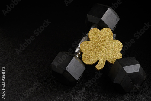Fototapeta Naklejka Na Ścianę i Meble -  Holiday fitness and Happy St. Patick’s Day, gold glitter shamrock shape with a pair of dumbbells on a black background
