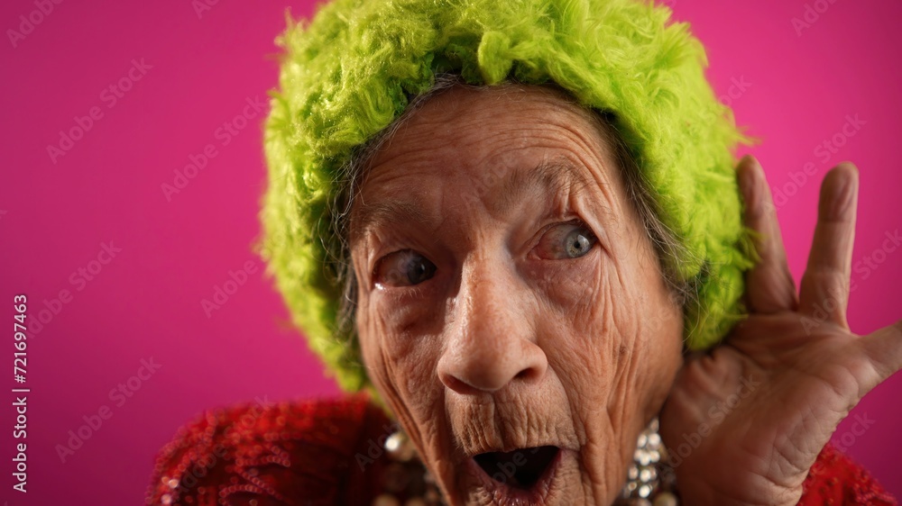 Funny fisheye view of smiling happy crazy grandmother with no teeth and wrinkled skin puts hand to ear to listen to a secret in isolated on pink background studio