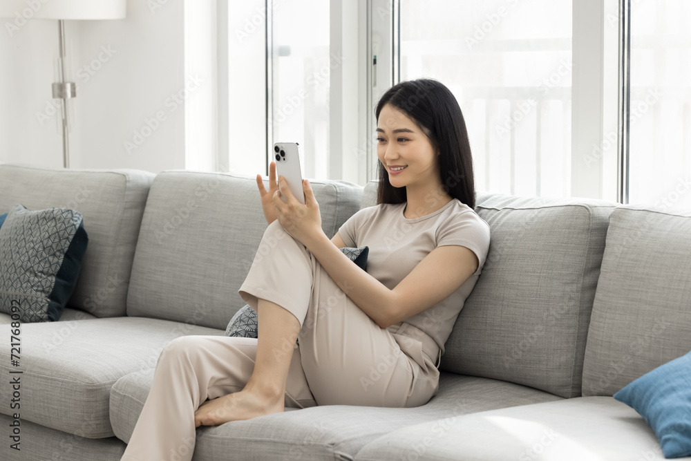 Happy beautiful young Asian blogger woman recording self video, talking selfie, talking on conference call on smartphone, using mobile phone on home sofa, holding gadget with knee support Happy beautiful young Asian blogger woman recording self video, talking selfie, talking on conference call on smartphone, using mobile phone on home sofa, holding gadget with knee support