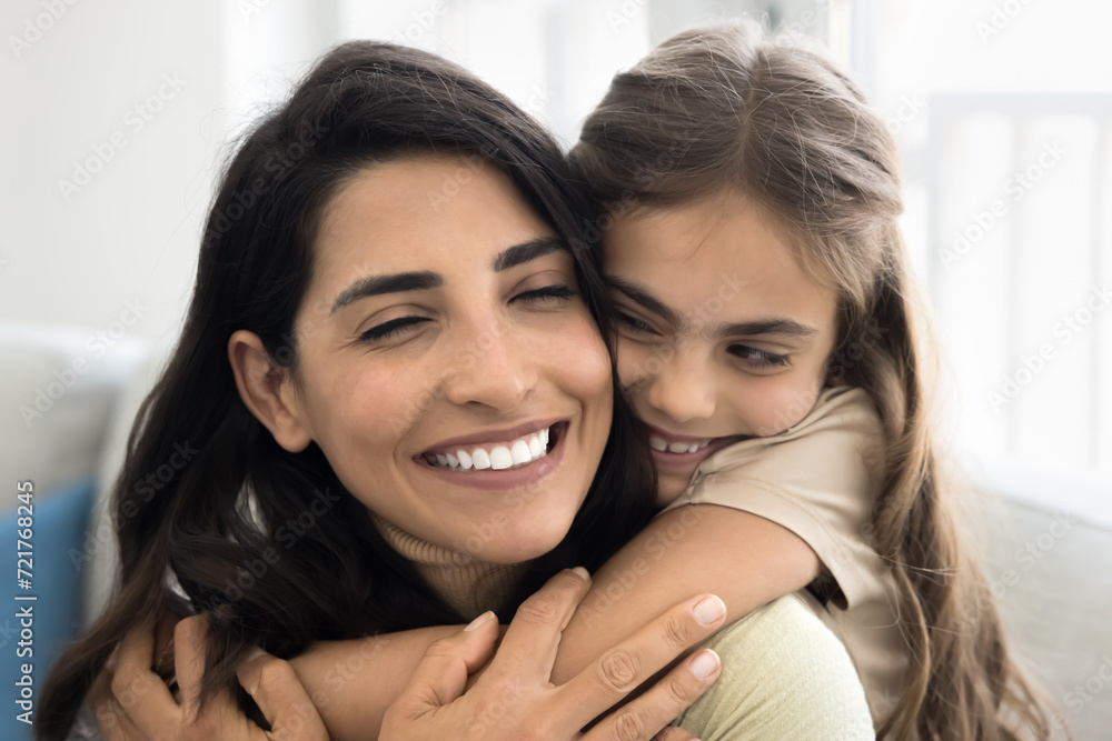 Cheerful cute daughter kid embracing happy mom from behind. Beautiful Latin mother piggybacking child with closed eyes and toothy smile, enjoying good close family relationship, motherhood Cheerful cute daughter kid embracing happy mom from behind. Beautiful Latin mother piggybacking child with closed eyes and toothy smile, enjoying good close family relationship, motherhood