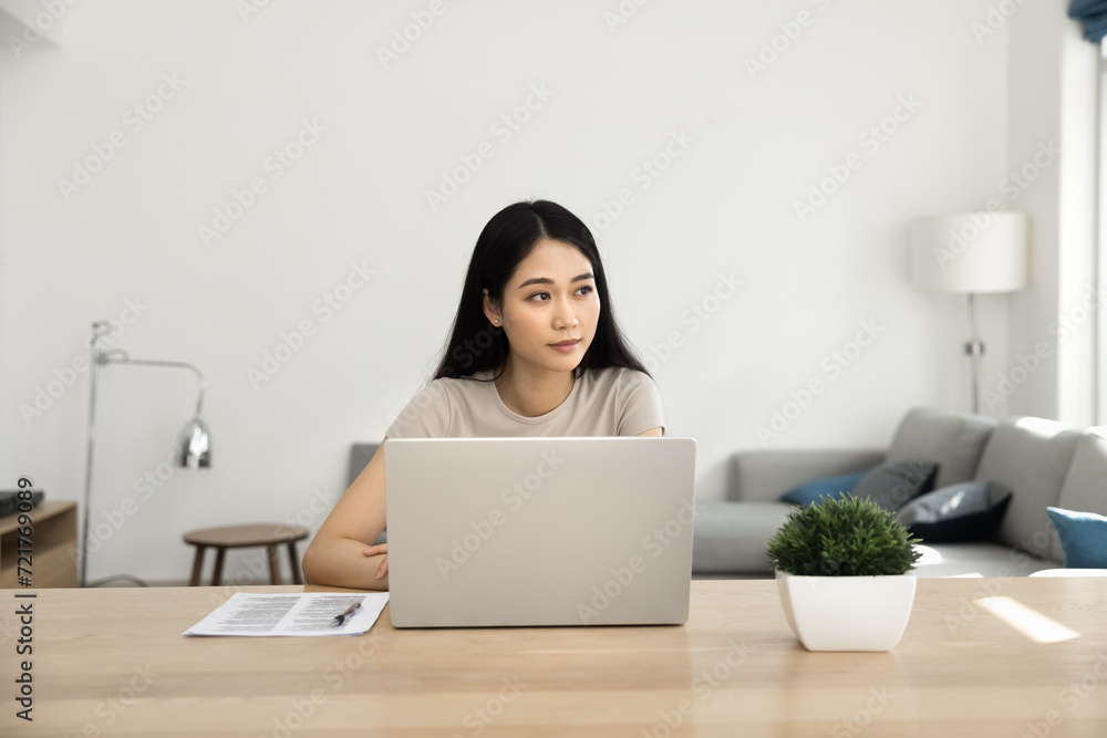Thoughtful young Asian freelance business woman sitting at laptop at home workplace, looking away, using Internet communication. Student girl working on research study at computer Thoughtful young Asian freelance business woman sitting at laptop at home workplace, looking away, using Internet communication. Student girl working on research study at computer