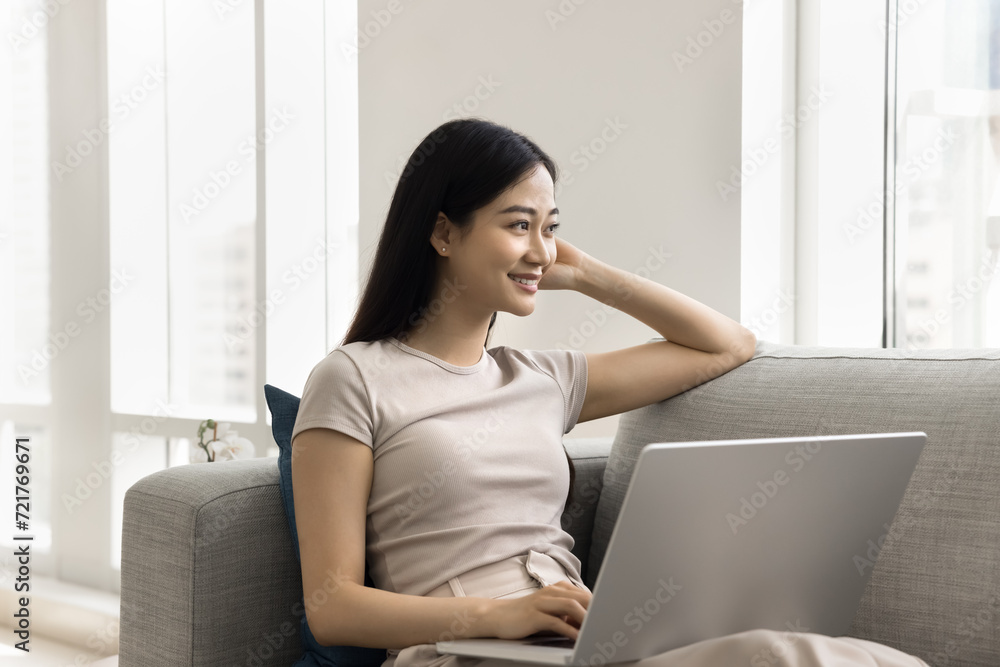 Happy thoughtful young Asian freelancer woman working at home, resting on couch with laptop, looking away, thinking on creative ideas, using wireless Internet technology, touching face Happy thoughtful young Asian freelancer woman working at home, resting on couch with laptop, looking away, thinking on creative ideas, using wireless Internet technology, touching face