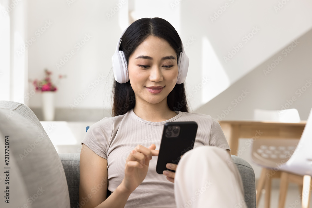 Happy pretty young Asian girl in headphones using smartphone, typing, setting online playlist, relaxing on comfortable couch at home, listening to music, enjoying leisure Happy pretty young Asian girl in headphones using smartphone, typing, setting online playlist, relaxing on comfortable couch at home, listening to music, enjoying leisure